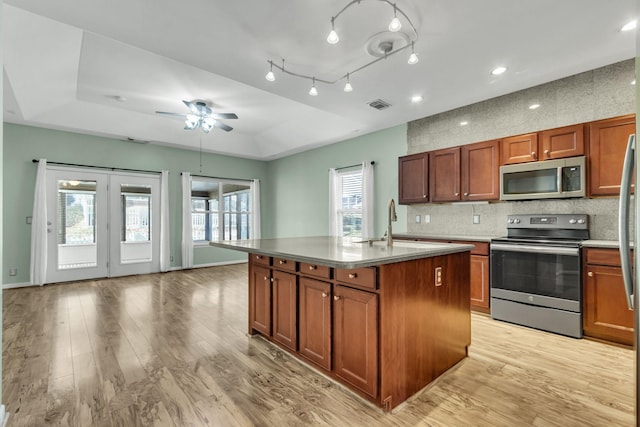 kitchen with ceiling fan, stainless steel appliances, a raised ceiling, light hardwood / wood-style floors, and a center island with sink