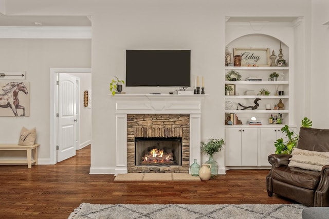 living room featuring built in features, ornamental molding, a stone fireplace, and dark wood-type flooring