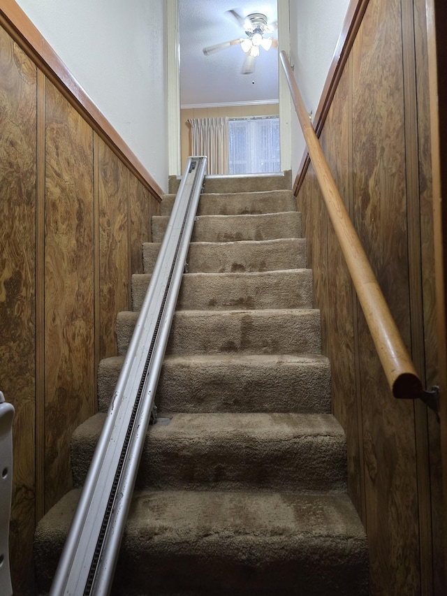 stairs featuring ceiling fan, ornamental molding, and wood walls