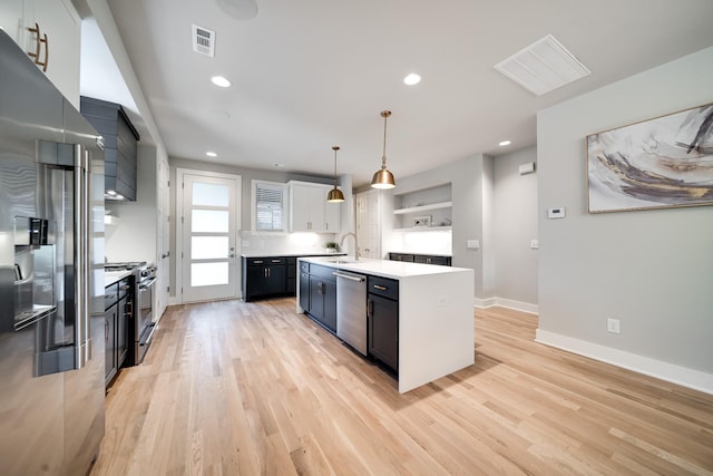 kitchen with white cabinetry, hanging light fixtures, stainless steel appliances, a kitchen island with sink, and light wood-type flooring