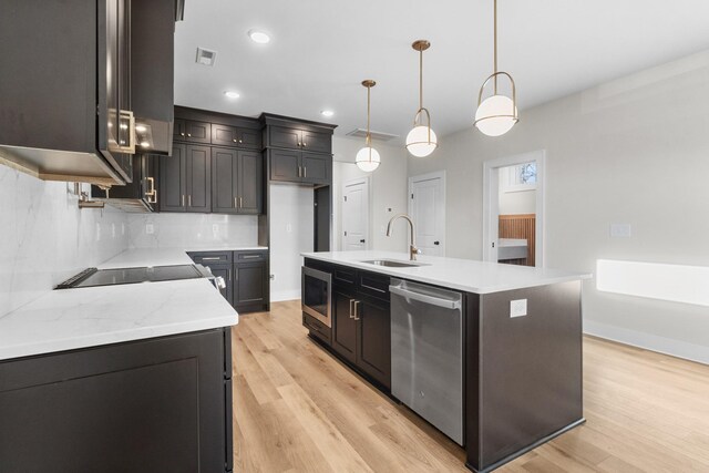 kitchen with sink, light wood-type flooring, an island with sink, appliances with stainless steel finishes, and decorative light fixtures