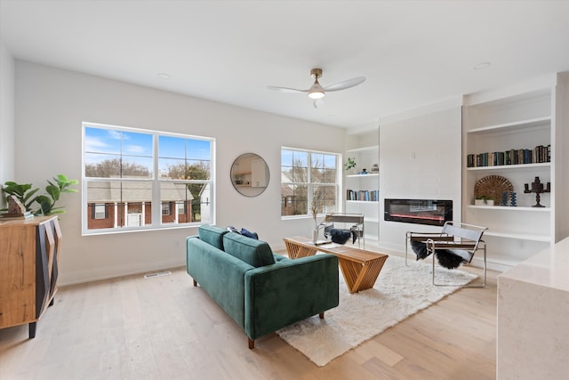 living area featuring built in features, visible vents, light wood-style floors, a glass covered fireplace, and ceiling fan