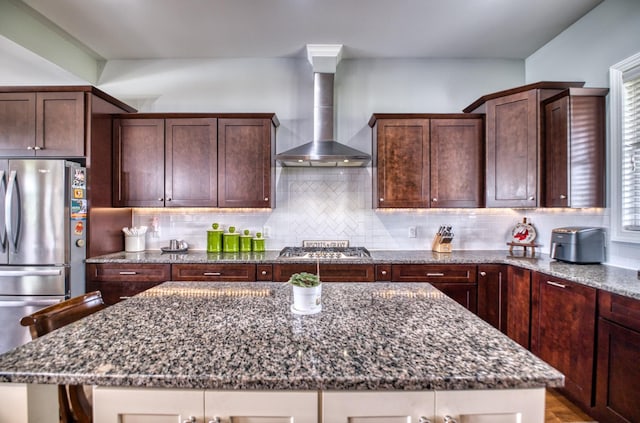 kitchen featuring wall chimney range hood, backsplash, dark stone countertops, a kitchen island, and appliances with stainless steel finishes