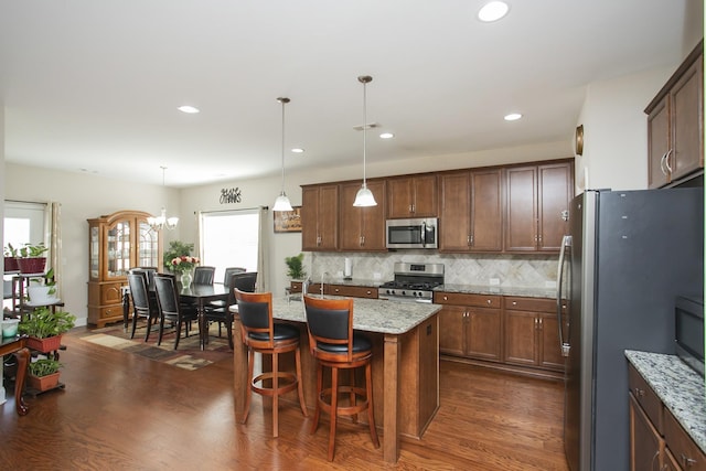 kitchen featuring light stone countertops, dark wood-type flooring, hanging light fixtures, an island with sink, and appliances with stainless steel finishes