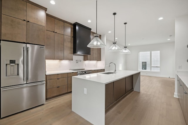 kitchen featuring appliances with stainless steel finishes, light wood-type flooring, a kitchen island with sink, wall chimney range hood, and hanging light fixtures