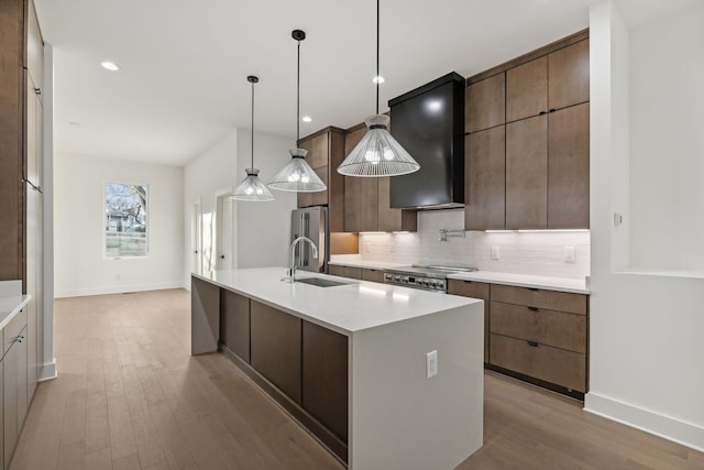 kitchen with wall chimney exhaust hood, sink, a center island with sink, light hardwood / wood-style floors, and hanging light fixtures