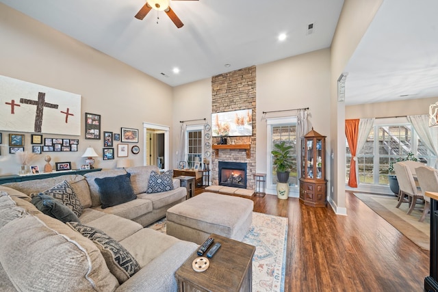 living room with ceiling fan, a stone fireplace, wood-type flooring, and a towering ceiling