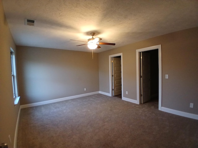 unfurnished bedroom with dark carpet, ceiling fan, a textured ceiling, a walk in closet, and multiple windows