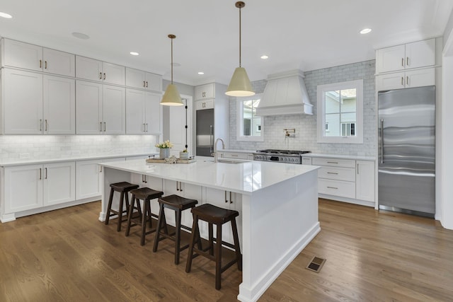 kitchen featuring dark hardwood / wood-style flooring, premium range hood, a kitchen island with sink, white cabinets, and appliances with stainless steel finishes