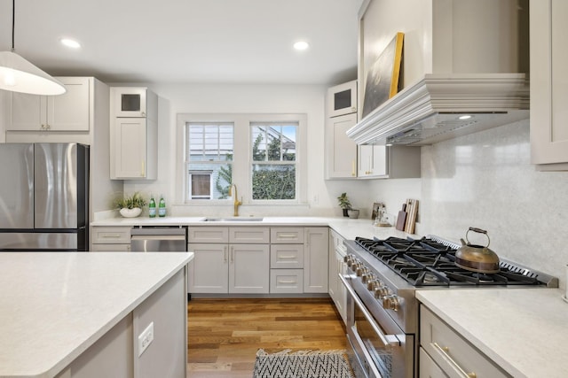 kitchen featuring sink, custom exhaust hood, hanging light fixtures, stainless steel appliances, and light hardwood / wood-style flooring