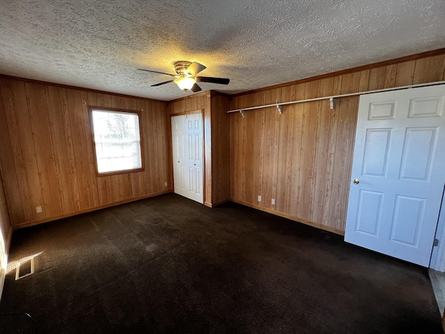 unfurnished bedroom featuring a textured ceiling, dark carpet, ceiling fan, and wood walls