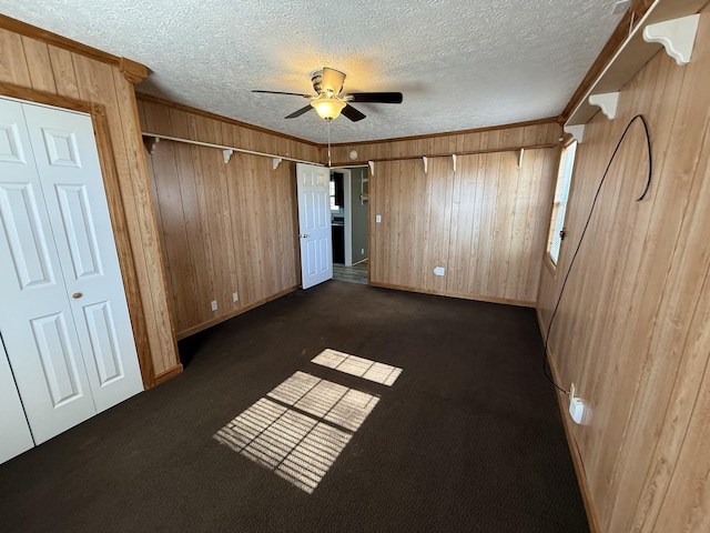 unfurnished bedroom featuring ceiling fan, a textured ceiling, wooden walls, dark carpet, and ornamental molding