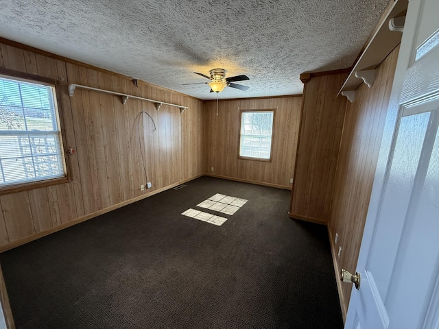 unfurnished room featuring ceiling fan, a healthy amount of sunlight, wooden walls, and dark carpet