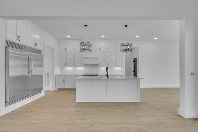 kitchen featuring sink, pendant lighting, stainless steel built in fridge, white cabinets, and an island with sink