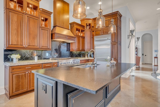 kitchen featuring appliances with stainless steel finishes, sink, custom range hood, decorative light fixtures, and a kitchen island with sink