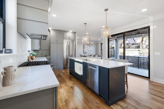 kitchen featuring sink, light hardwood / wood-style flooring, appliances with stainless steel finishes, hanging light fixtures, and an island with sink