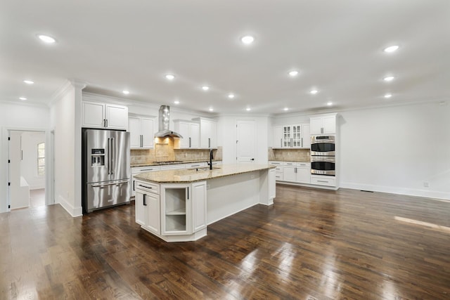 kitchen featuring dark wood-type flooring, a center island with sink, wall chimney range hood, appliances with stainless steel finishes, and white cabinetry