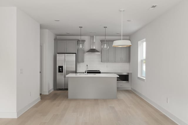 kitchen featuring gray cabinetry, wall chimney range hood, light wood-type flooring, an island with sink, and appliances with stainless steel finishes