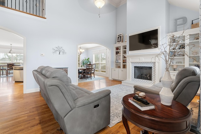 living room featuring light wood-type flooring, built in features, a fireplace, a towering ceiling, and ceiling fan with notable chandelier