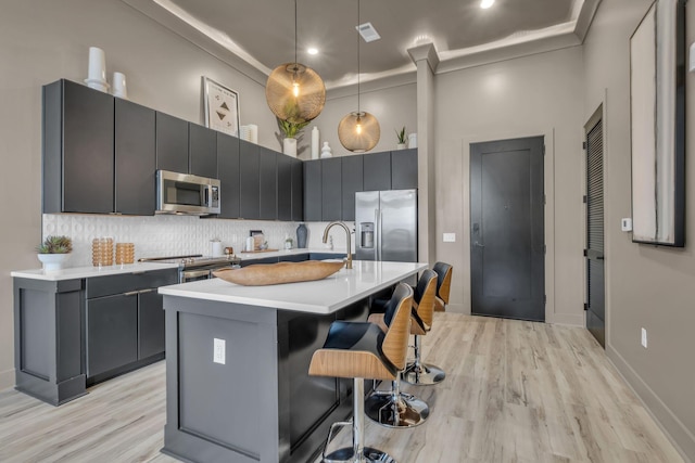 kitchen with crown molding, light wood-type flooring, an island with sink, appliances with stainless steel finishes, and decorative light fixtures