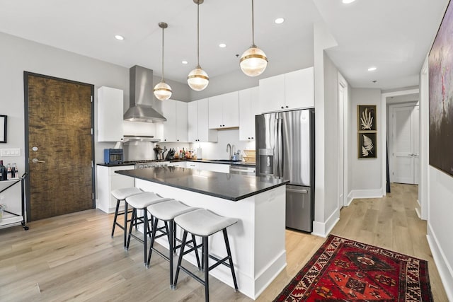 kitchen with light wood-type flooring, stainless steel appliances, wall chimney range hood, white cabinets, and hanging light fixtures