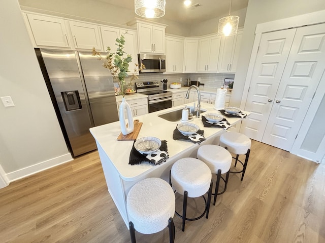 kitchen featuring a kitchen island with sink, sink, appliances with stainless steel finishes, decorative light fixtures, and white cabinetry