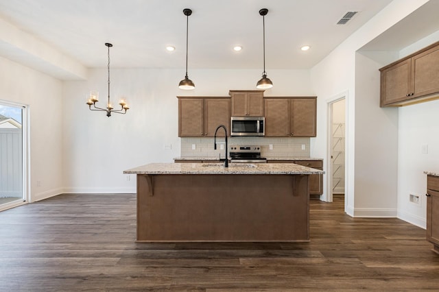 kitchen with stainless steel appliances, light stone counters, hanging light fixtures, and a kitchen island with sink