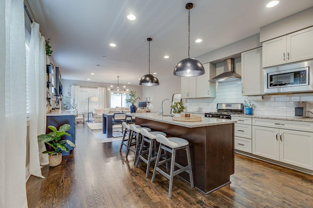 kitchen with appliances with stainless steel finishes, dark hardwood / wood-style flooring, wall chimney exhaust hood, a center island with sink, and white cabinetry