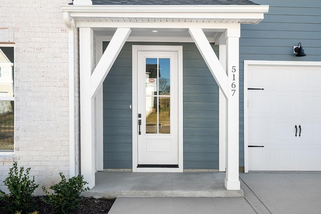 view of exterior entry with a garage, covered porch, a shingled roof, and brick siding