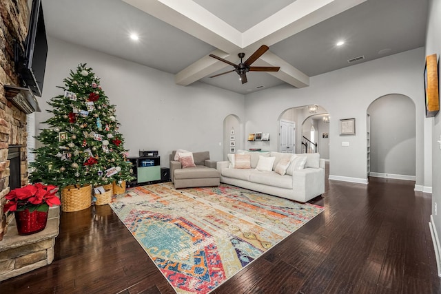 living room featuring ceiling fan, beam ceiling, a stone fireplace, and dark wood-type flooring