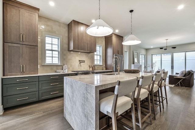 kitchen with light stone countertops, a kitchen island with sink, pendant lighting, wood-type flooring, and decorative backsplash