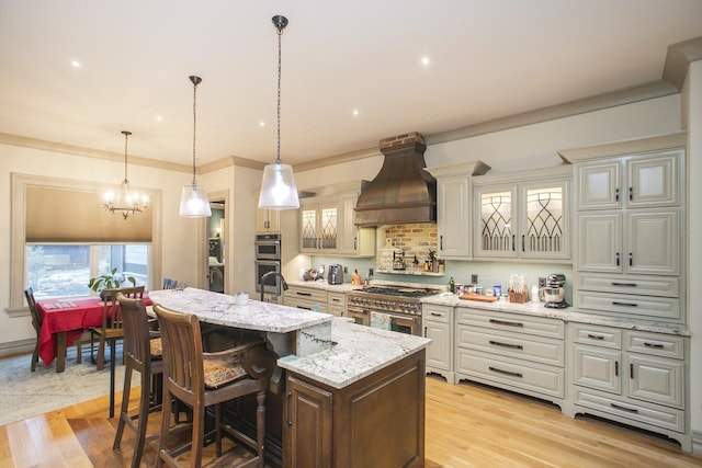 kitchen featuring a kitchen island with sink, light stone counters, light hardwood / wood-style floors, custom range hood, and decorative light fixtures