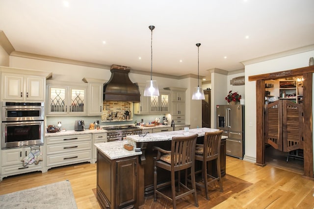 kitchen featuring premium range hood, high end appliances, light stone counters, hanging light fixtures, and a kitchen island with sink