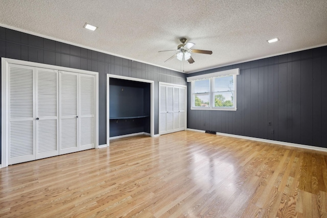 unfurnished bedroom featuring light wood-type flooring, a textured ceiling, two closets, ceiling fan, and wooden walls