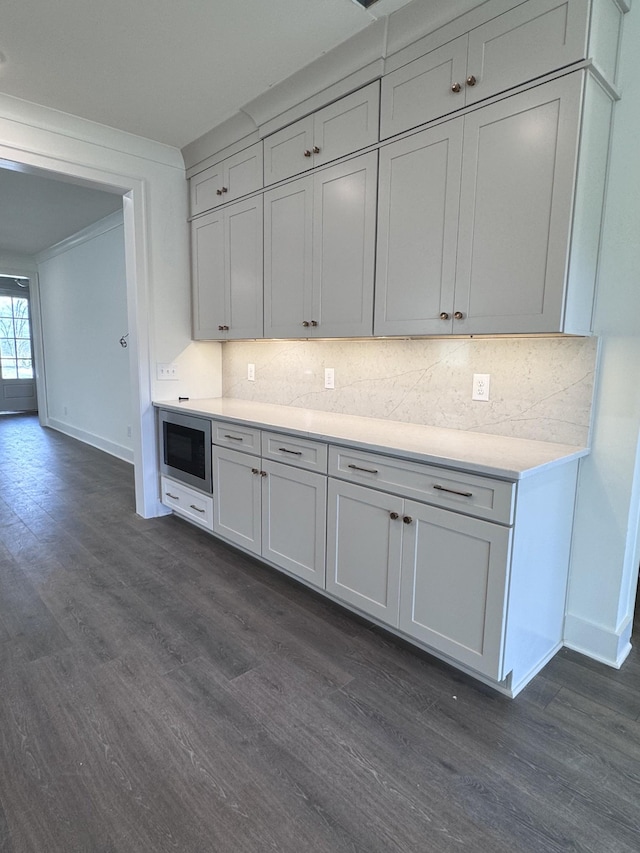 kitchen featuring dark wood-type flooring, stainless steel microwave, light countertops, decorative backsplash, and baseboards