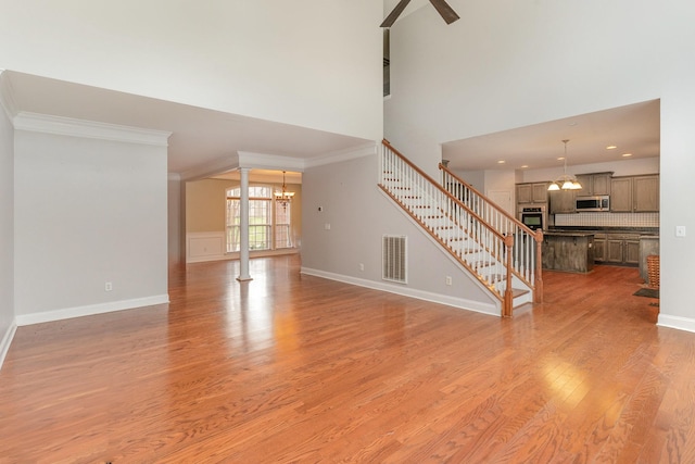 unfurnished living room featuring decorative columns, crown molding, light wood-type flooring, a towering ceiling, and ceiling fan with notable chandelier