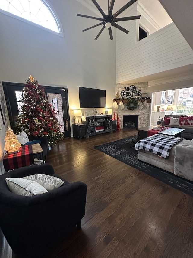 living room featuring a stone fireplace, ceiling fan, a towering ceiling, and dark wood-type flooring