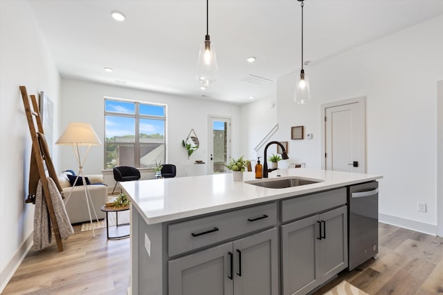 kitchen featuring stainless steel dishwasher, sink, decorative light fixtures, light hardwood / wood-style floors, and an island with sink