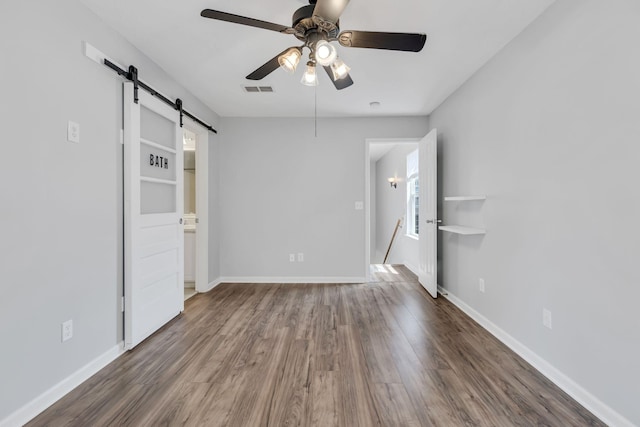 unfurnished bedroom featuring a barn door, ceiling fan, ensuite bathroom, and dark wood-type flooring