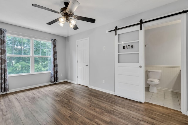 unfurnished bedroom featuring hardwood / wood-style floors, ceiling fan, a barn door, and ensuite bathroom