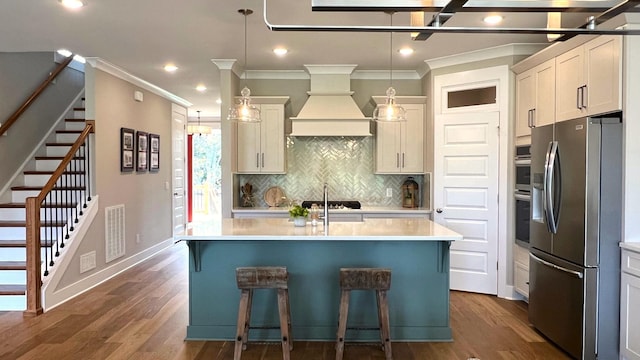 kitchen with hanging light fixtures, dark wood-type flooring, premium range hood, stainless steel fridge, and an island with sink