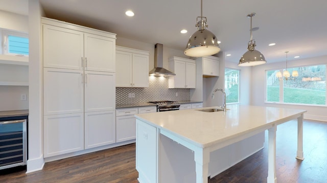 kitchen with sink, wall chimney range hood, wine cooler, hanging light fixtures, and an island with sink