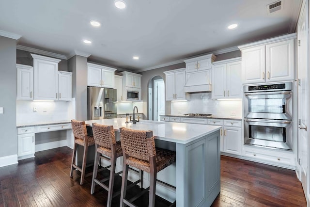 kitchen with a kitchen island with sink, sink, custom range hood, white cabinetry, and stainless steel appliances