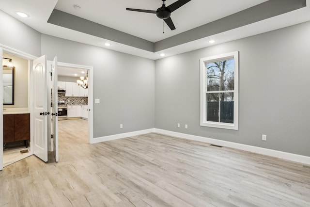 empty room with ceiling fan with notable chandelier, light wood-type flooring, and a tray ceiling