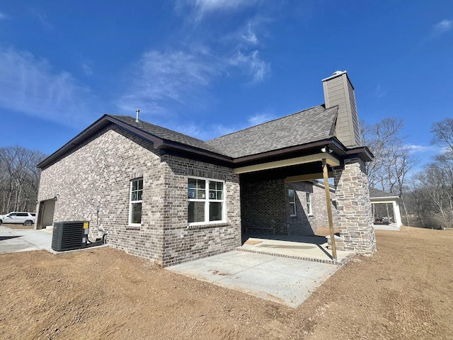 exterior space with a shingled roof, a patio, a chimney, central AC, and brick siding