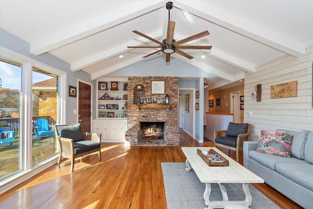 living room featuring built in shelves, ceiling fan, a brick fireplace, vaulted ceiling with beams, and wood-type flooring