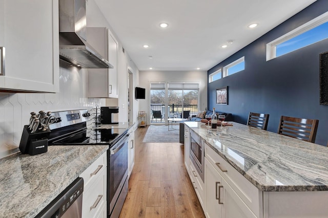 kitchen with white cabinets, a breakfast bar area, wall chimney exhaust hood, light wood-type flooring, and stainless steel appliances