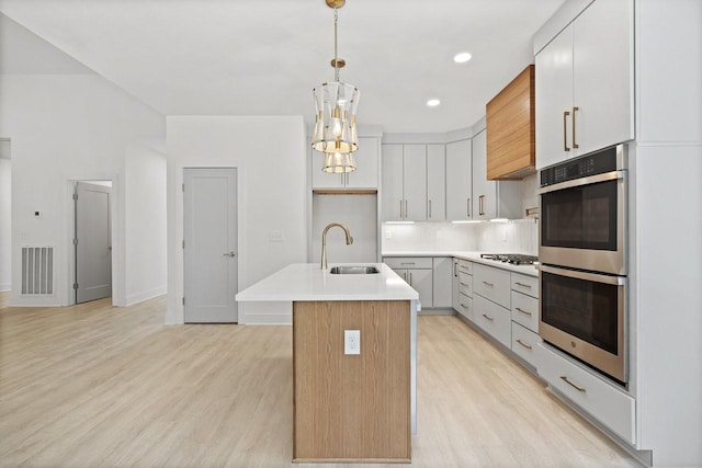 kitchen with a kitchen island with sink, sink, light hardwood / wood-style flooring, appliances with stainless steel finishes, and decorative light fixtures