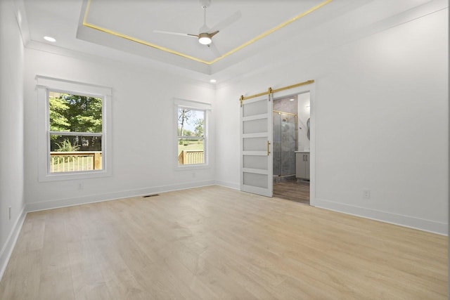 unfurnished bedroom featuring ensuite bath, ceiling fan, a barn door, light wood-type flooring, and a tray ceiling