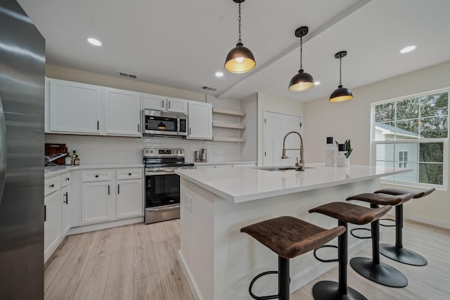 kitchen with a kitchen island with sink, hanging light fixtures, light hardwood / wood-style floors, white cabinetry, and stainless steel appliances